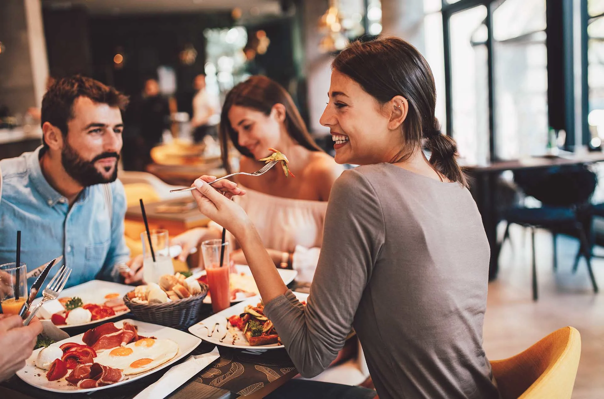 Four people sit around a restaurant table enjoying food and drinks. One woman in the foreground smiles while holding a fork. The atmosphere is bright and casual, with natural light and modern decor.